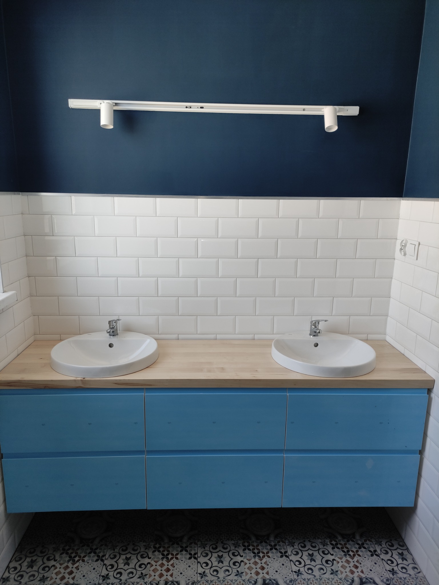 Wykończona łazienka z dwiema umywalkami nablatowymi, białe płytki typu metro, szafka w odcieniach błękitu, drewniany blat, ozdobna podłoga i nowoczesne oświetlenie punktowe.