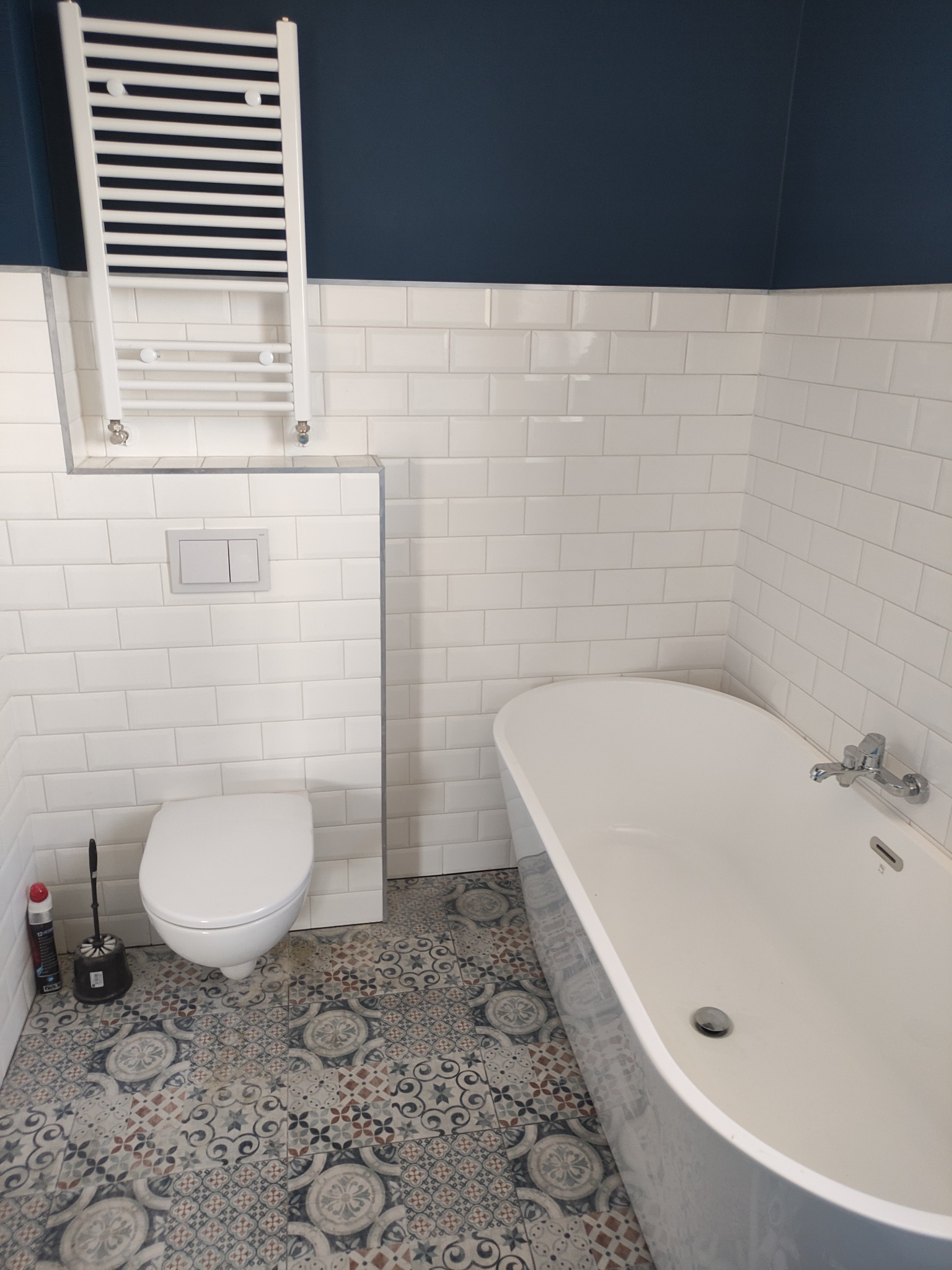 Wykończona łazienka z białą armaturą, grzejnikiem drabinkowym i dekoracyjną podłogą w stylu retro, ściany wyłożone białymi, prostokątnymi płytkami.