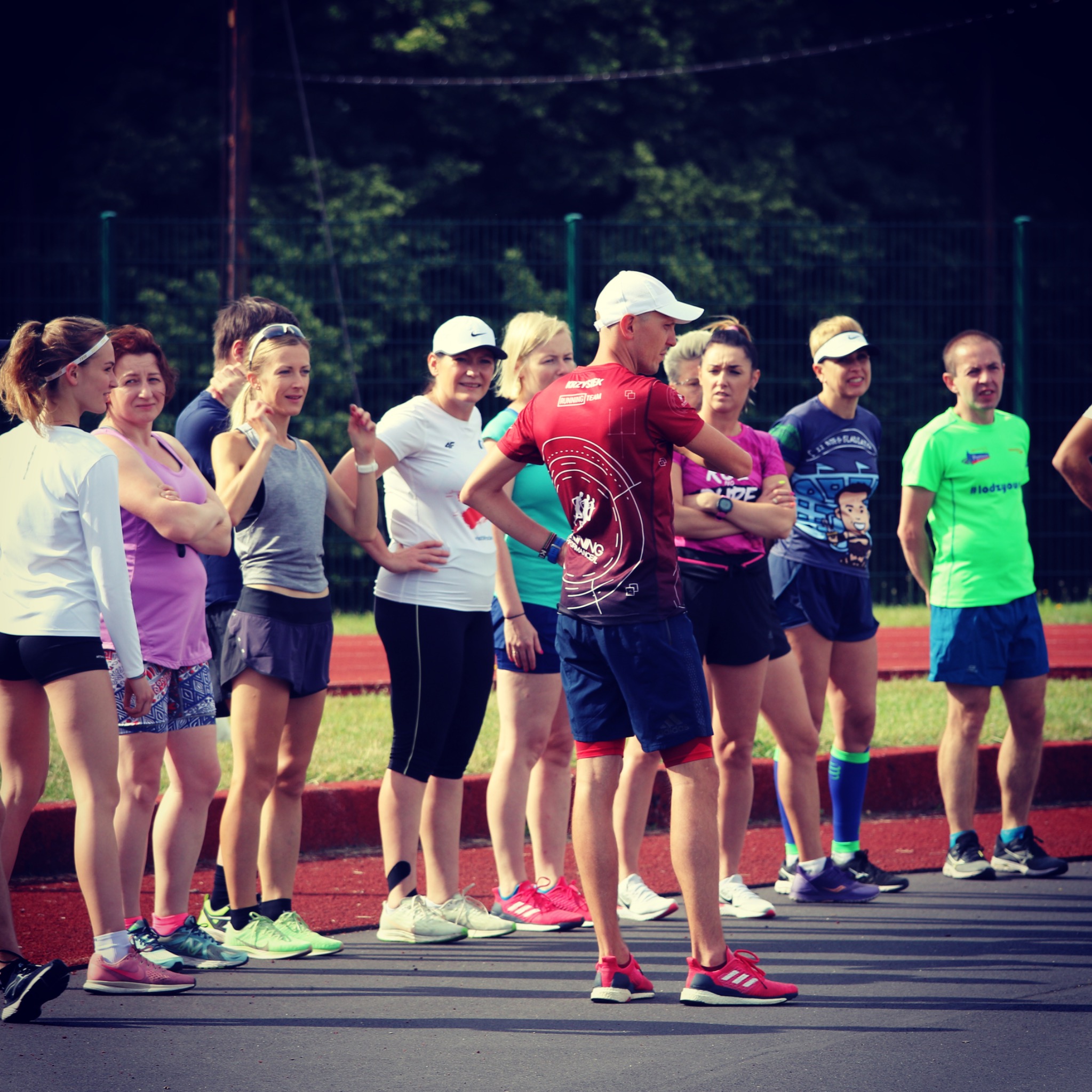 Grupa osób w strojach sportowych, w tym mężczyzna w czerwonej koszulce z napisem 'Running Team', stojąca na bieżni, prawdopodobnie słuchająca instrukcji przed treningiem.