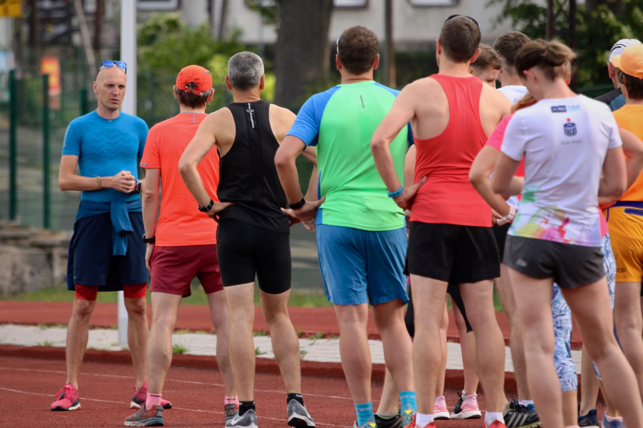 Grupa biegaczy na bieżni, z przodu mężczyzna w niebieskiej koszulce i okularach na głowie, reszta osób stoi tyłem, w różnokolorowych strojach sportowych, ręce oparte na biodrach.