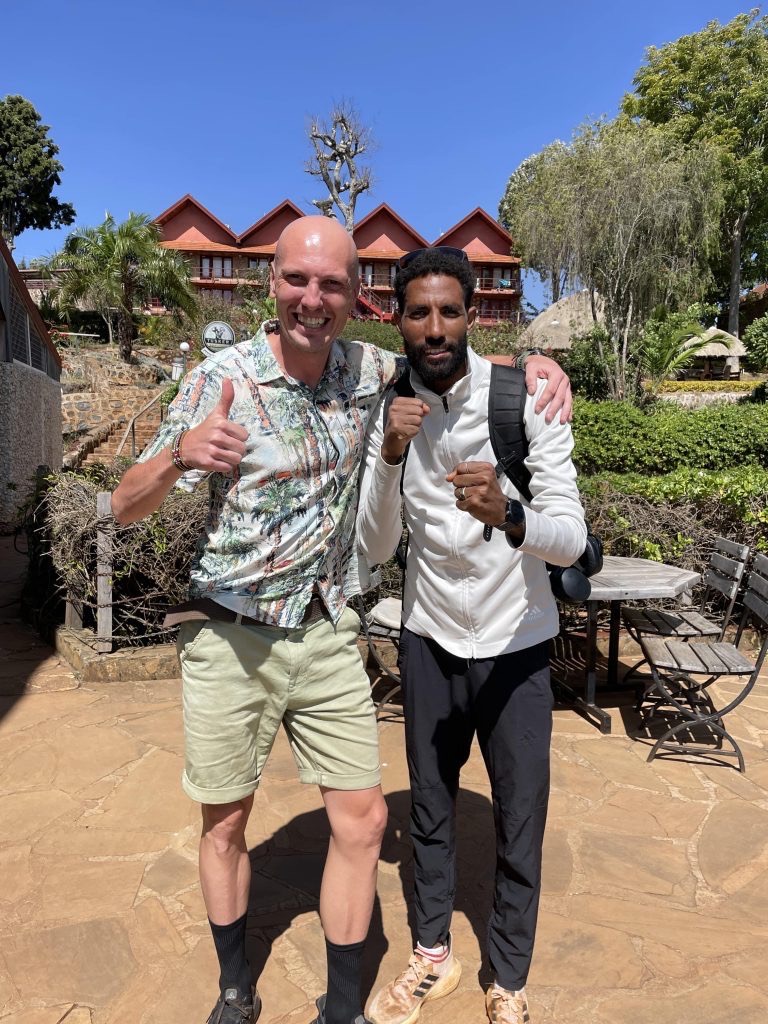 Uśmiechnięty mężczyzna z kciukiem w górę obejmuje boksera w pozycji gotowej do walki na tle egzotycznego krajobrazu z budynkami o czerwonych dachach i bujną roślinnością.