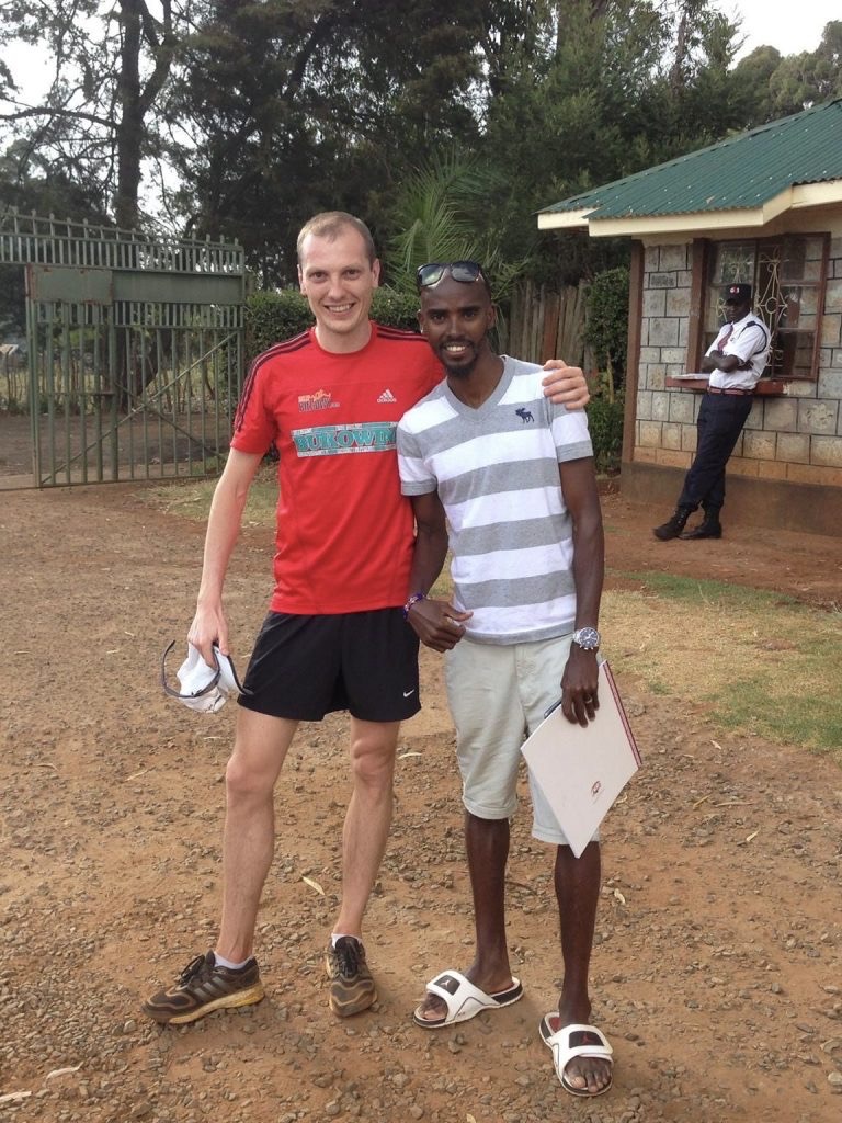 Dwóch mężczyzn pozuje do zdjęcia na tle budynku z ochroniarzem; jeden w czerwonej koszulce i butach do biegania, drugi w koszulce w paski i sandałach, trzymający notatnik.