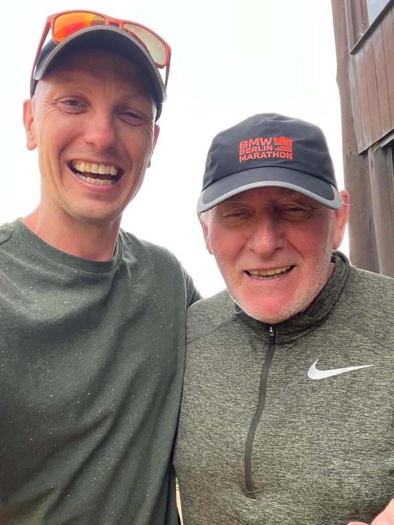 Dwóch uśmiechniętych mężczyzn, jeden w czapce BMW Berlin Marathon, drugi w okularach przeciwsłonecznych na czapce, pozuje razem, prawdopodobnie po treningu biegowym.