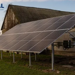 Instalacja fotowoltaiczna na gruncie z widocznym inwerterem i starą stodołą w tle, logo firmy ATUAL w lewym g&oacute;rnym rogu.