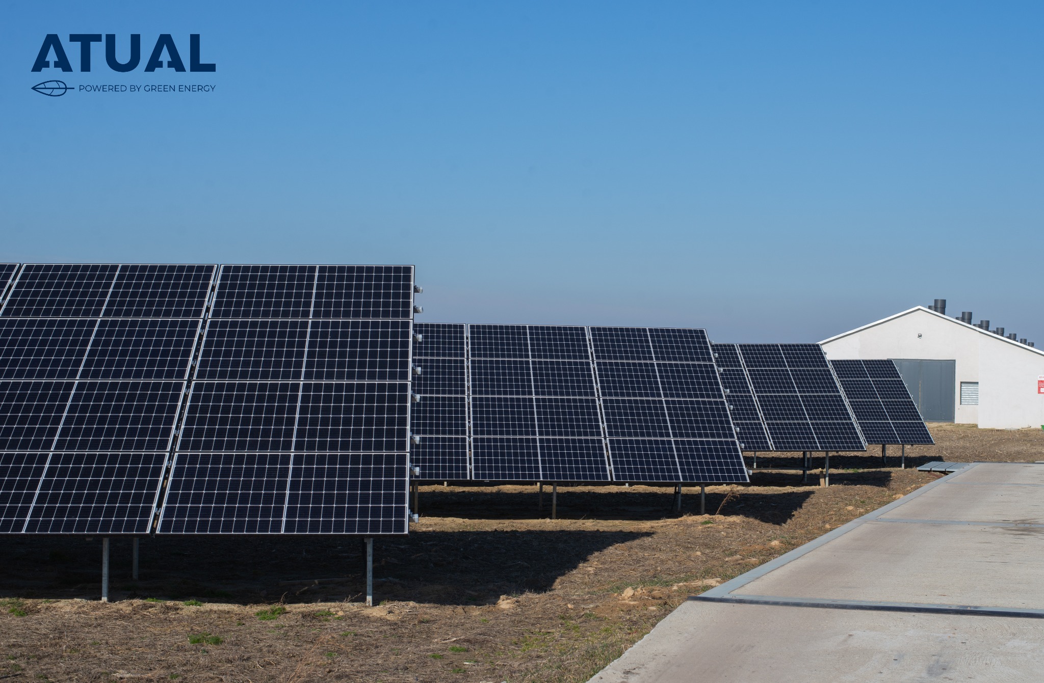 Rząd paneli słonecznych na polnej ziemi, z białym budynkiem w tle i logo firmy w lewym górnym rogu.