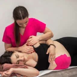 Kobieta w różowym uniformie wykonuje masaż na plecach pacjentki leżącej na stole; widoczne różowe plastry kinesiotaping na biodrze pacjentki.