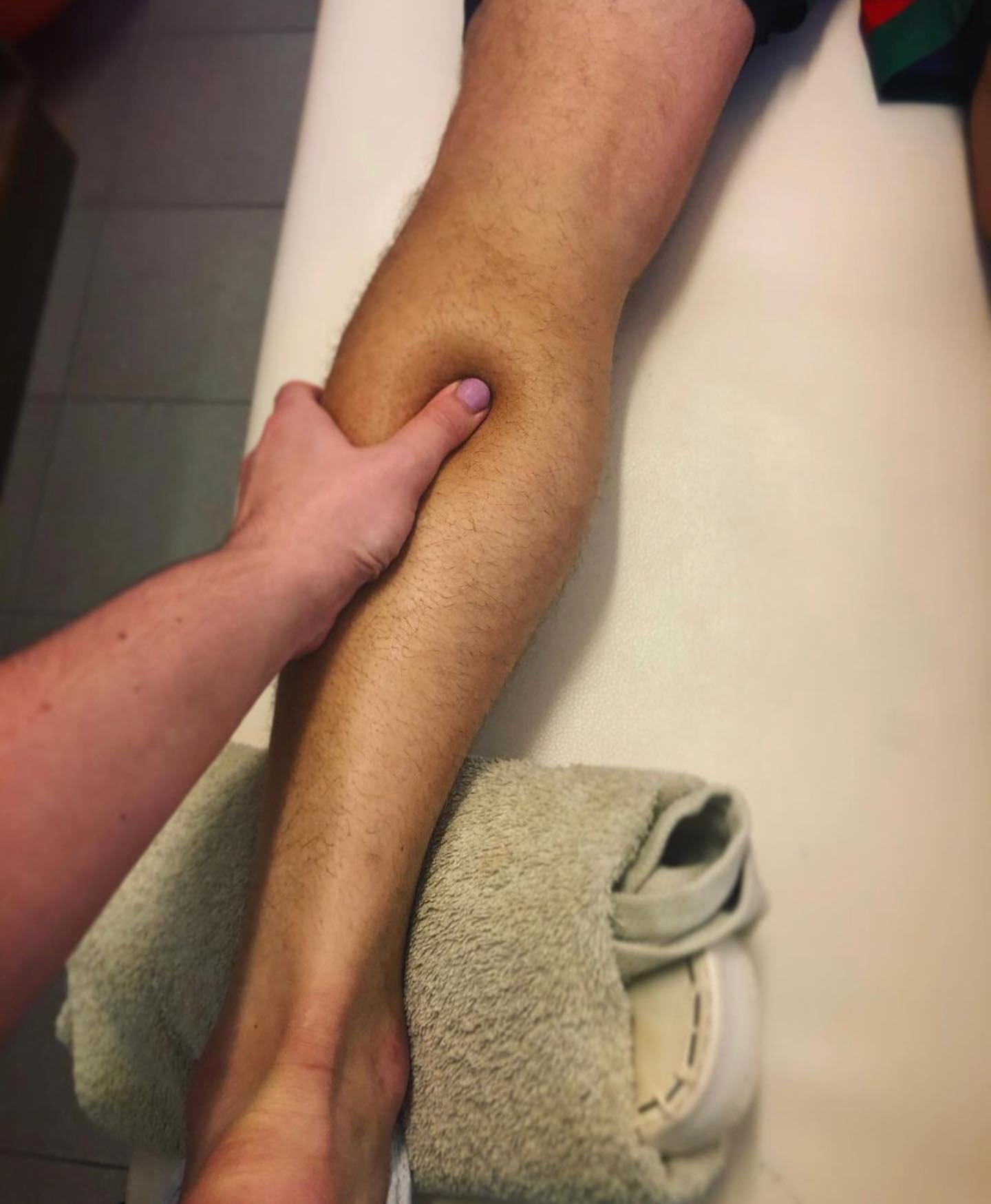 Masaż łydki pacjenta na leżance, dłoń z fioletowym lakierem na paznokciach uciska mięsień, stopa oparta na ręczniku.