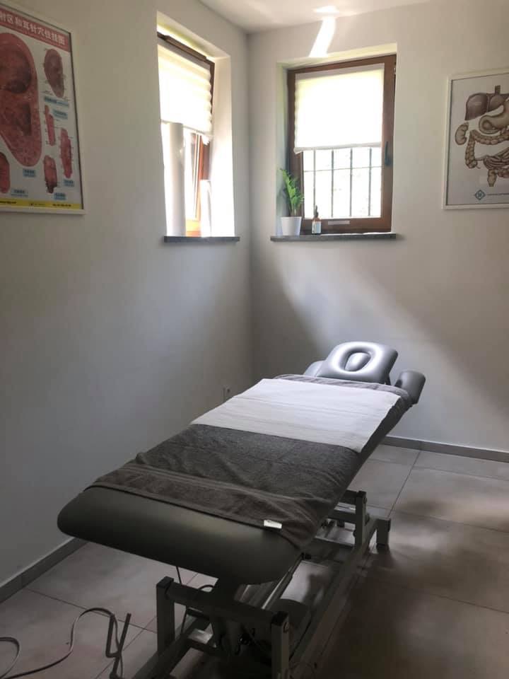 Poczekalnia z kozetką do zabiegów, dwa okna z roletami, plakaty anatomiczne na ścianach, szary ręcznik i białe prześcieradło na leżance.