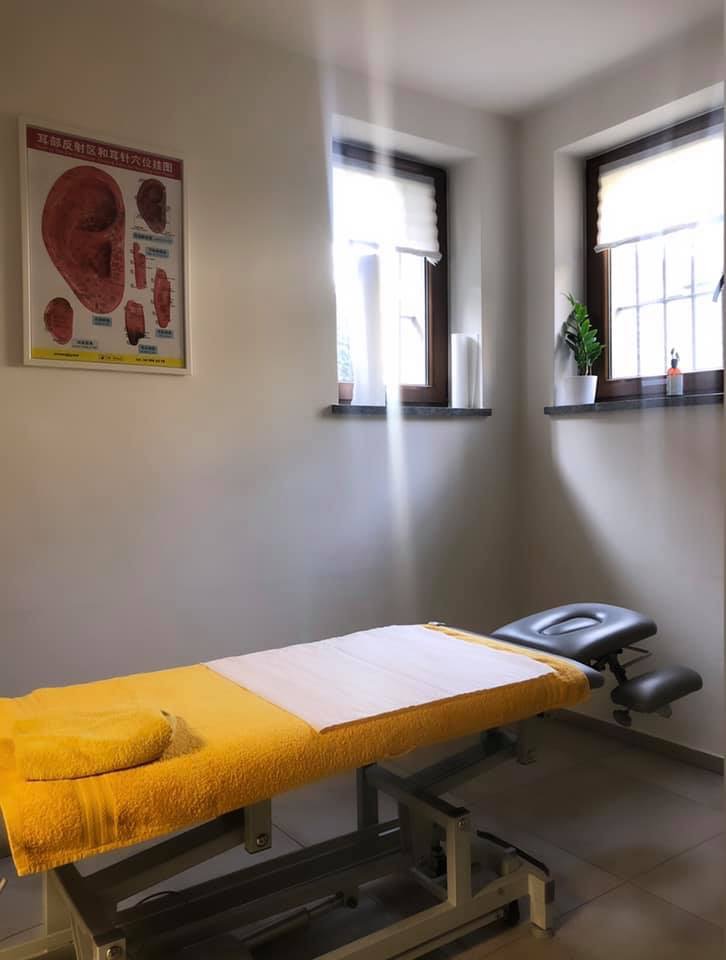 Pokój zabiegowy z regulowanym łóżkiem pokrytym żółtym ręcznikiem i białą podkładką, plakat z mapą ucha na ścianie, jasne światło wpadające przez okna.