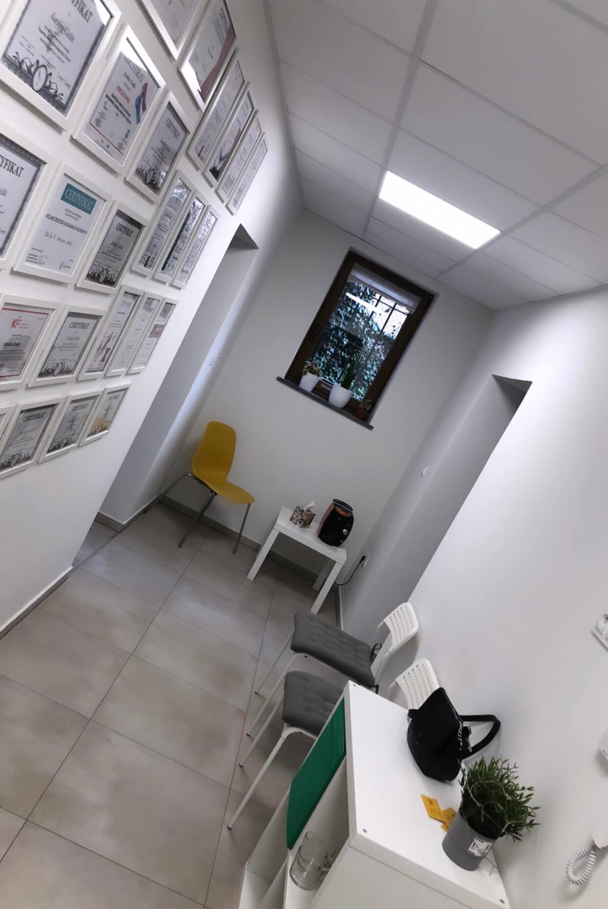 Jasne wnętrze poczekalni z widoczną ścianą z certyfikatami w ramkach, żółtym krzesłem, białym stolikiem z czajnikiem i oknem z roślinami, a także z szarymi krzesłami dla pacjentów