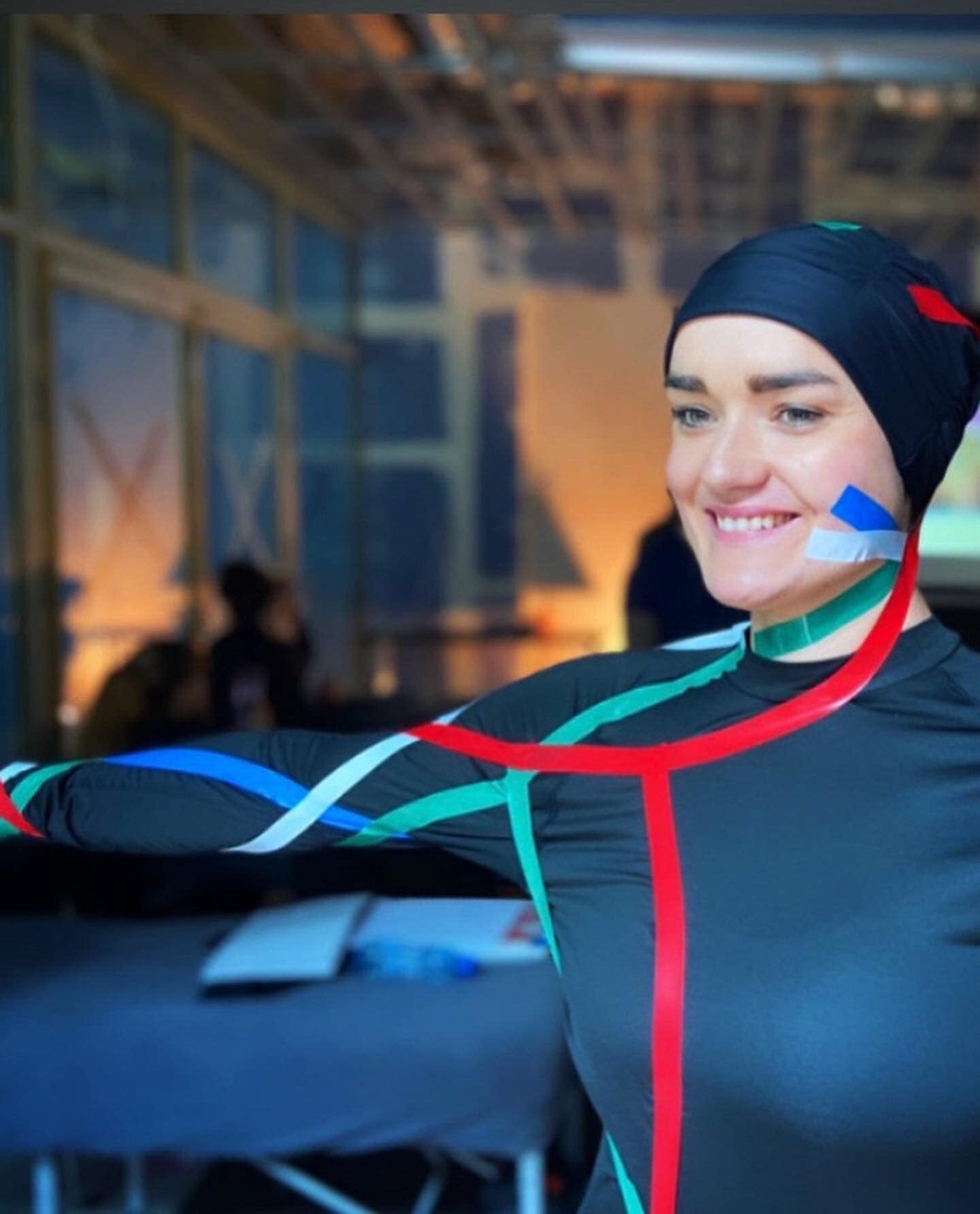 Uśmiechnięta kobieta w czarnym stroju z kolorowymi taśmami i czepku, przygotowywana do analizy ruchu w laboratorium biomechanicznym.