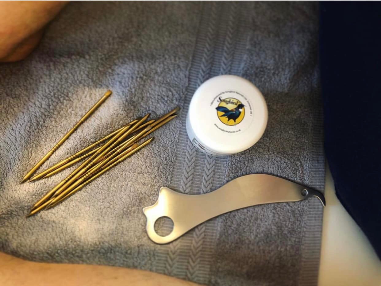 Zestaw narzędzi do terapii manualnej: złote igły, krem z logo Songbird i metalowy skrobak Gua Sha na szarym ręczniku.