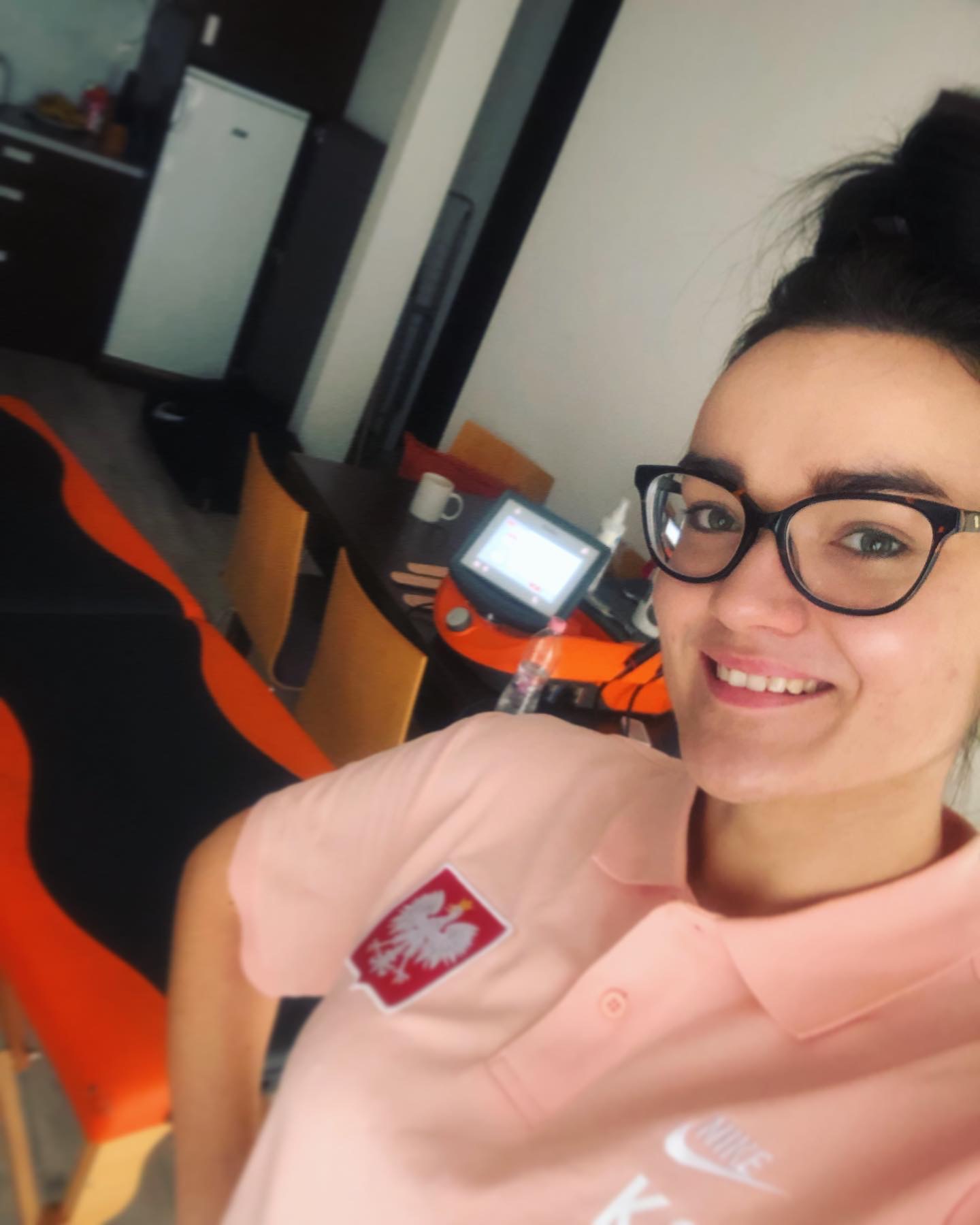 Uśmiechnięta kobieta w okularach i koszulce z godłem Polski, pozująca w pomieszczeniu z fotelem gamingowym i urządzeniem do fizjoterapii.