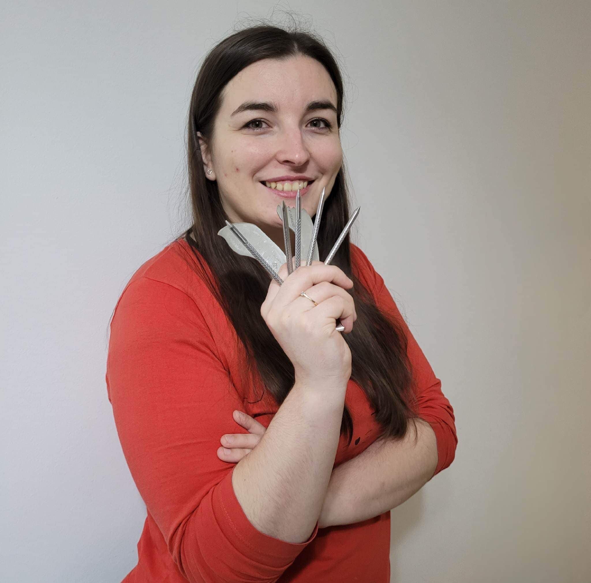 Uśmiechnięta kobieta w czerwonej koszulce trzyma w dłoni zestaw metalowych narzędzi do zabiegów medycyny alternatywnej, prezentując je na tle jasnej ściany.
