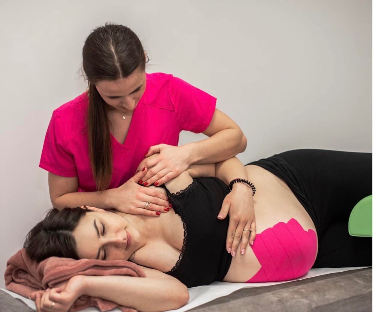 Kobieta w różowym uniformie wykonuje masaż na plecach pacjentki leżącej na stole; widoczne różowe plastry kinesiotaping na biodrze pacjentki.