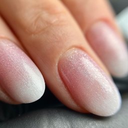 Atelier Piękna Sylwia Gajda - Zbliżenie na dłoń z delikatnym, połyskującym manicure ombre w odcieniach różu i bieli, na czarnym tle.