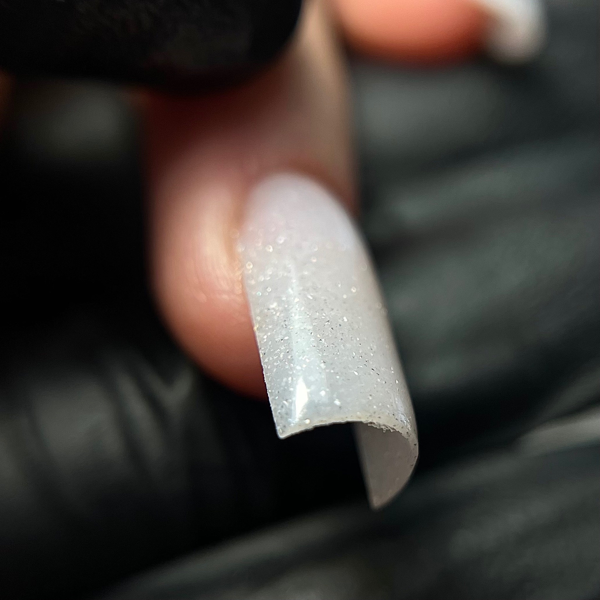 Powiększone ujęcie przezroczystego, błyszczącego tipsa z opiłowaną krawędzią, trzymanego w palcach w czarnych rękawiczkach.