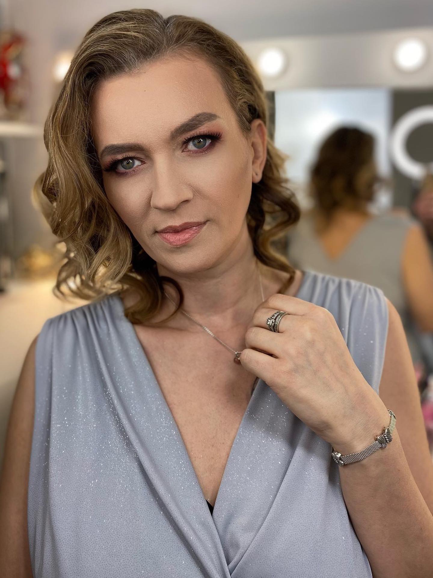 Portret kobiety z delikatnym, różowym cieniem na powiekach, subtelnym konturowaniem i błyszczącym pudrem na twarzy, w tle widać oświetlenie typowe dla salonu kosmetycznego.