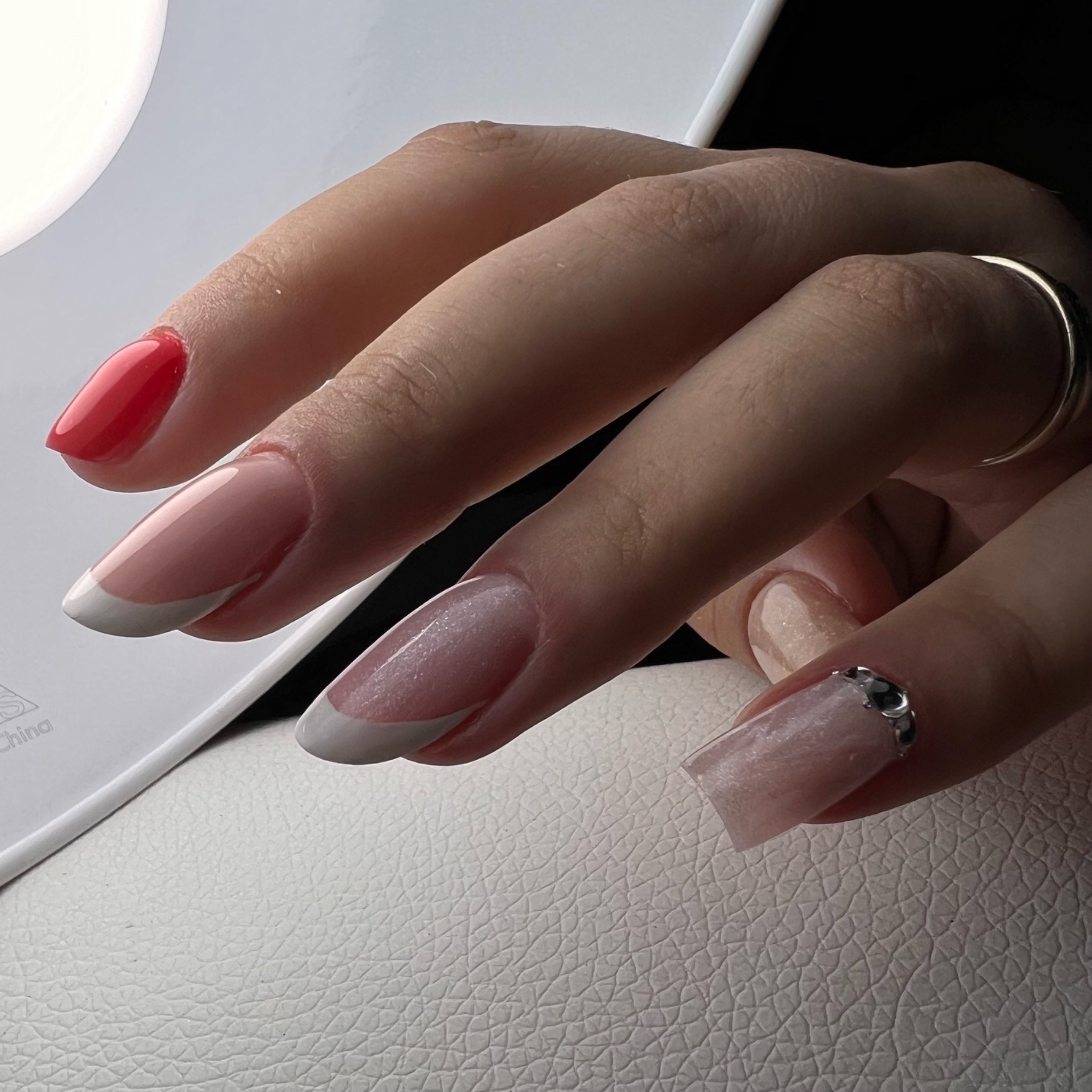 Dłoń z manicure hybrydowym: czerwony lakier na jednym paznokciu, klasyczny french na dwóch, brokatowy róż z ozdobnymi kamyczkami na jednym, pierścionek na palcu wskazującym, białe skórzane tło.