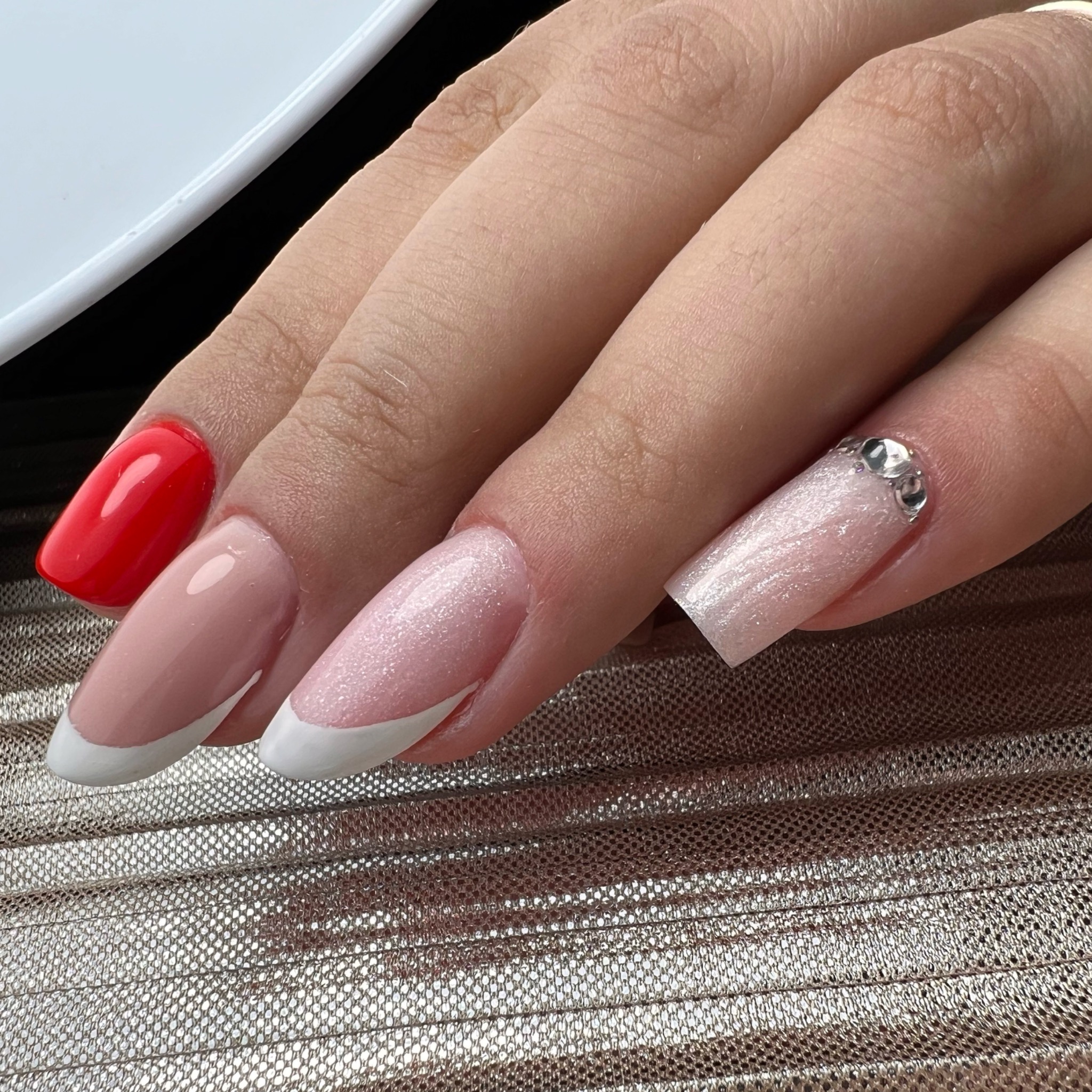 Dłoń z różnymi stylizacjami paznokci: czerwony, french manicure i brokatowy z cyrkoniami, na złotym, teksturowanym tle.