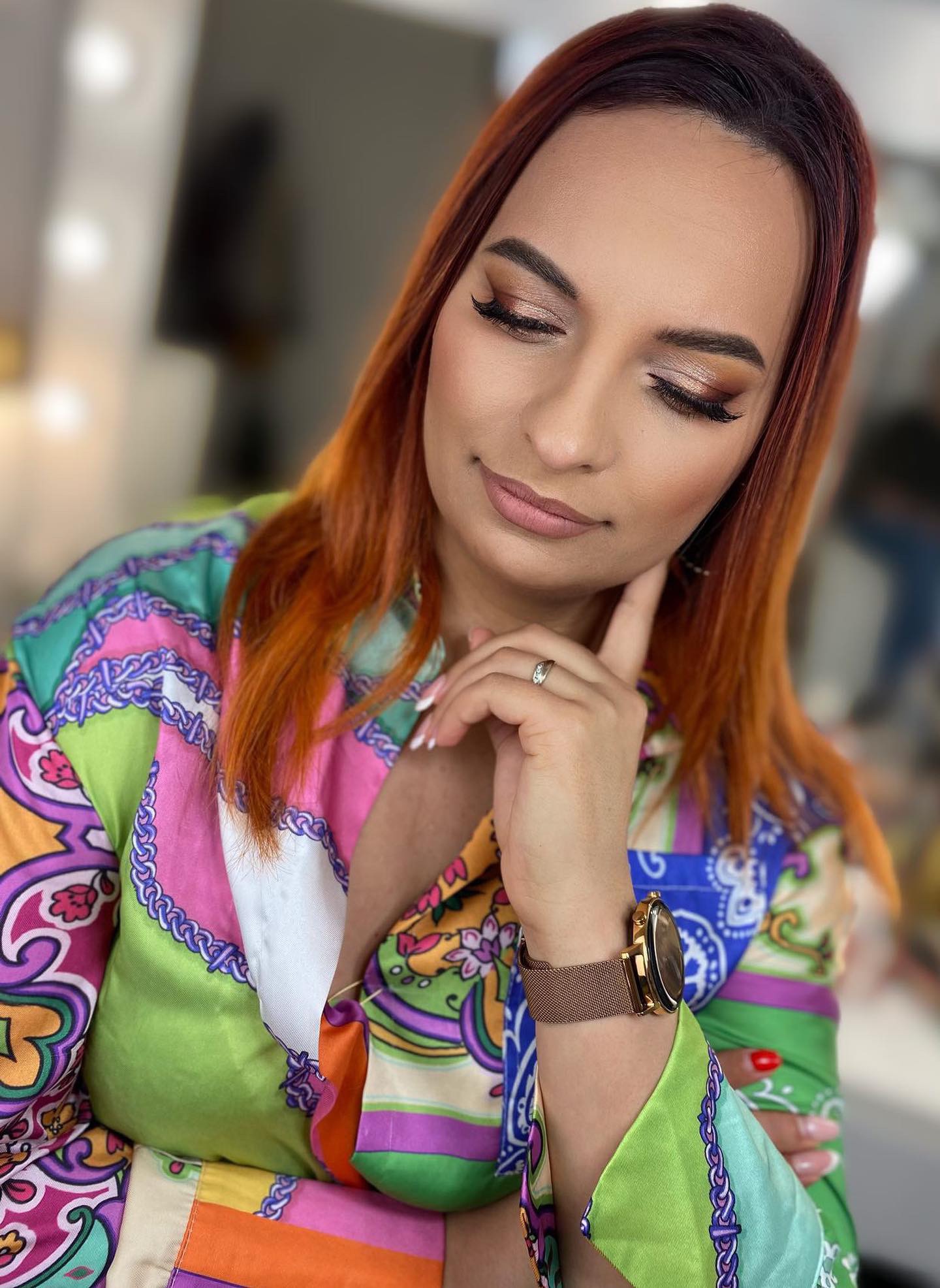 Portret kobiety z rudymi włosami i profesjonalnym, błyszczącym makijażem oczu w odcieniach brązu, z długimi, gęstymi rzęsami, ubrana w kolorową koszulę w stylu retro, pozująca z ręką pod brodą...