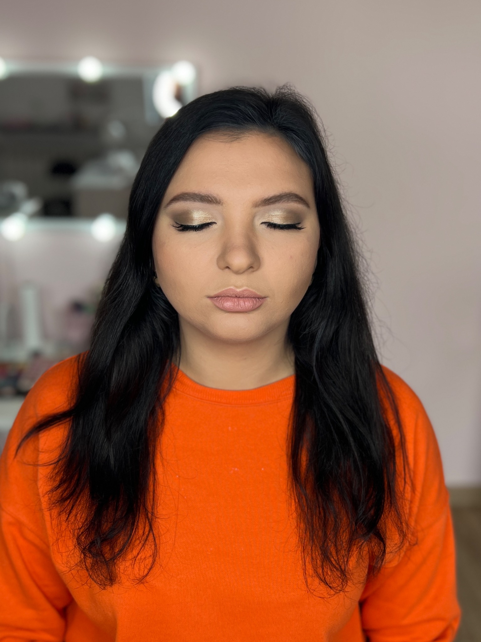 Kobieta z ciemnymi włosami i zamkniętymi oczami prezentuje delikatny, złoty makijaż oczu z subtelną kreską i naturalny makijaż ust w studio z oświetleniem.