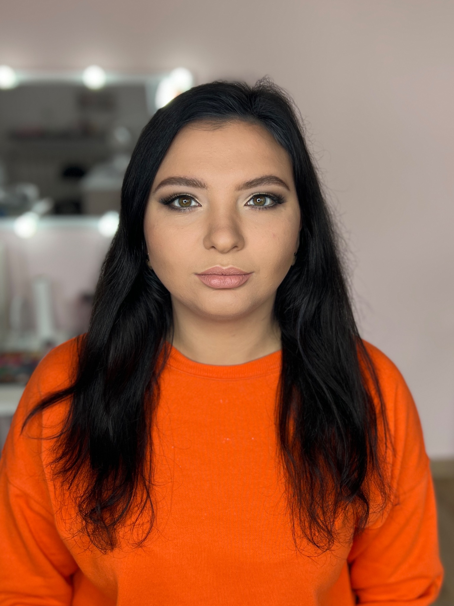 Portret kobiety z delikatnym makijażem w Strzyżowie, z uwydatnionymi oczami i naturalnymi ustami. Uwagę zwraca świeża cera i ciemne, proste włosy opadające na pomarańczową bluzę.