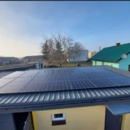 Instalacja fotowoltaiczna na dachu budynku gospodarczego z blachy trapezowej, w tle dom z zielonym dachem i drzewo bez liści.