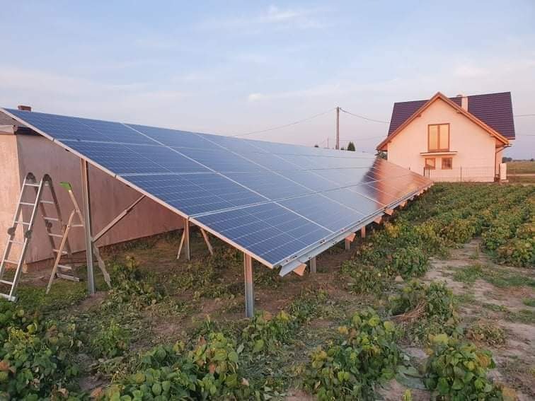 Instalacja fotowoltaiczna na gruncie w wiejskim otoczeniu, widoczna drabina oparta o konstrukcję, dom jednorodzinny w tle.