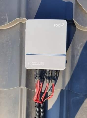 Szara skrzynka z logo FOX i napisem FIREFIGHTER SAFETY SWITCH, zamontowana na szarym dachu z widocznymi przewodami w czerwonej izolacji i czarnej peszli.