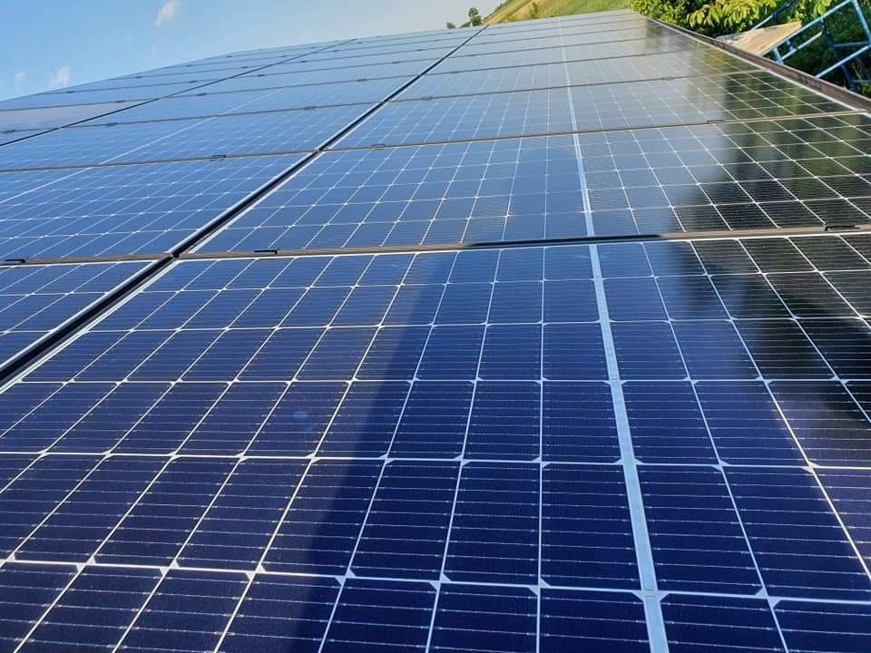 Niebieskie panele słoneczne na dachu, widok pod kątem, z widocznym fragmentem nieba i zieleni w tle.