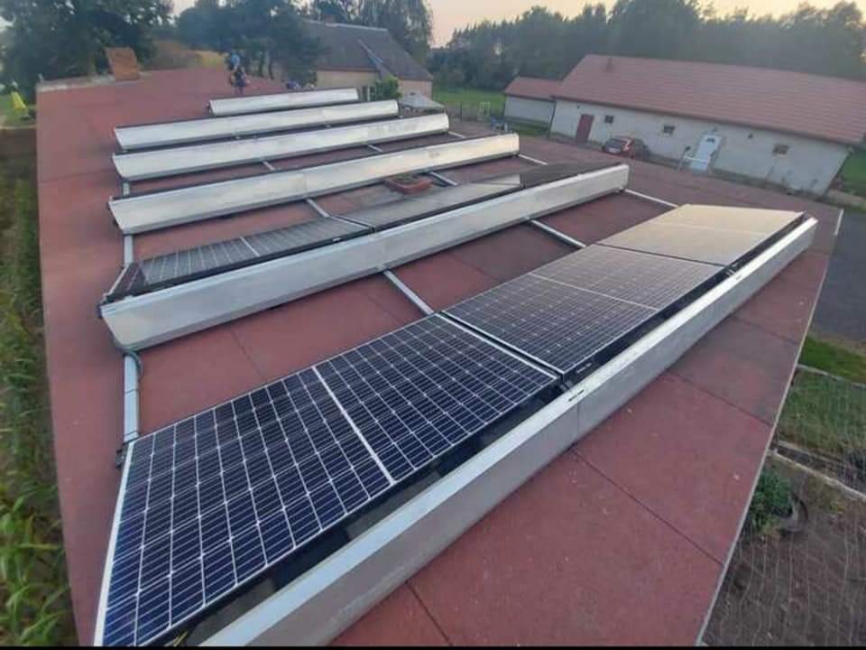 Rząd paneli słonecznych z aluminiową obudową zamontowanych na czerwonym dachu budynku gospodarczego, widoczne elementy montażowe i konstrukcja wsporcza.