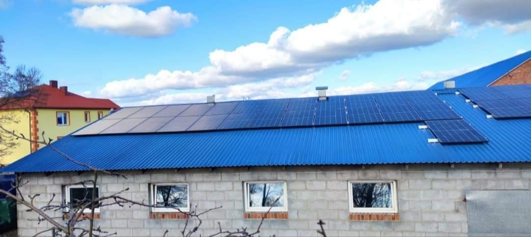 Instalacja paneli słonecznych na niebieskim, blaszany dachu budynku gospodarczego z szarego pustaka, widok z dołu w słoneczny dzień.