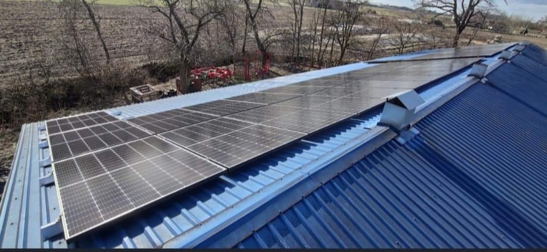 Instalacja paneli fotowoltaicznych na niebieskim, blaszanym dachu budynku gospodarczego, widok z góry na panele i okoliczne pole uprawne.