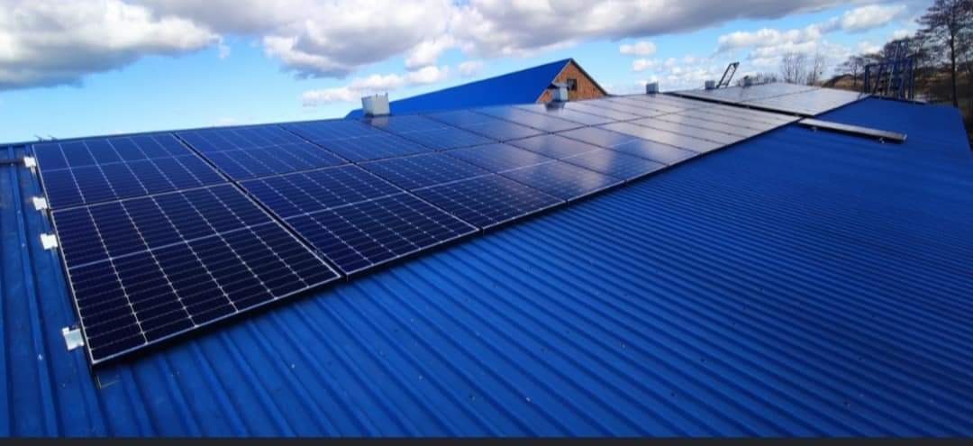Panele fotowoltaiczne zamontowane na niebieskim, metalowym dachu budynku z widocznym fragmentem konstrukcji wsporczej na tle błękitnego nieba z chmurami.