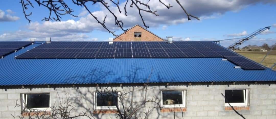 Instalacja paneli fotowoltaicznych na niebieskim, falistym dachu budynku gospodarczego z cegły, widoczne gałęzie drzew na pierwszym planie i konstrukcja wsporcza w tle.