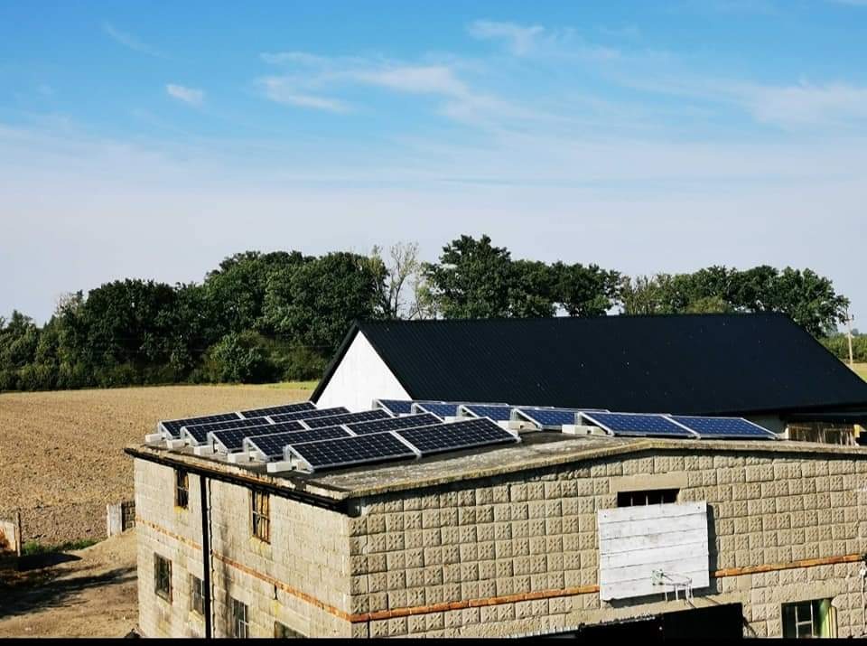 Panele słoneczne zamontowane na dachu budynku gospodarczego z cegły betonowej, w tle pole i las.