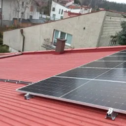 Panele słoneczne zamontowane na czerwonym dachu z blachy falistej, widoczne przewody prowadzone wzdłuż komina i dachu, w tle zabudowa jednorodzinna.