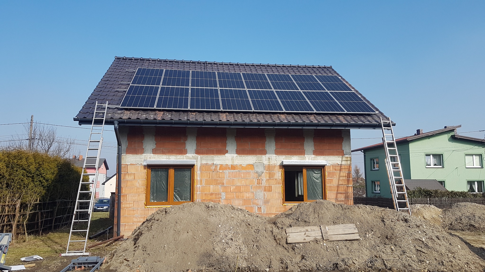 Surowy budynek z czerwonej cegły z zamontowanymi panelami słonecznymi na dachu, dwie drabiny opierają się o ściany, a przed budynkiem usypane są pryzmy ziemi.