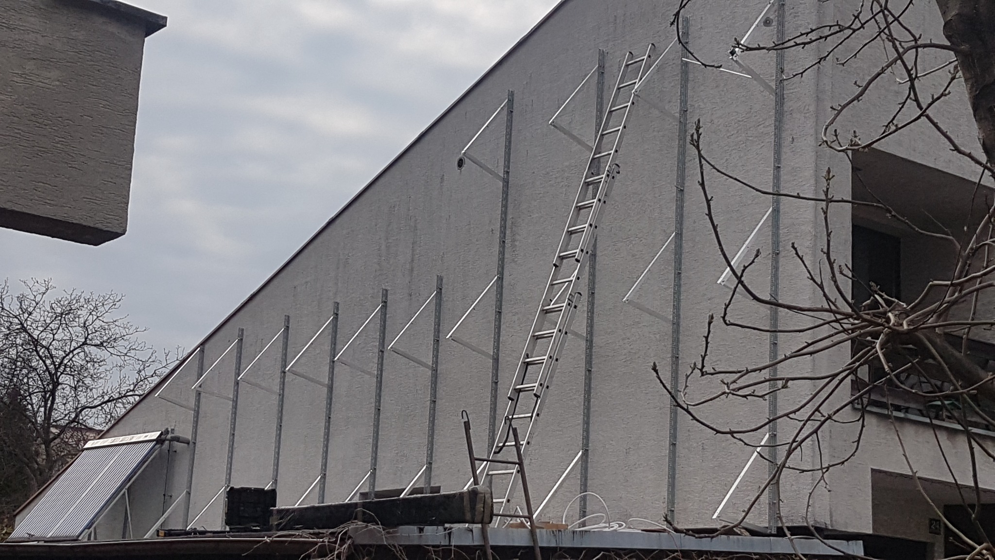 Montaż stelażu pod panele słoneczne na elewacji budynku, widoczna drabina oparta o ścianę i fragment paneli na dachu.
