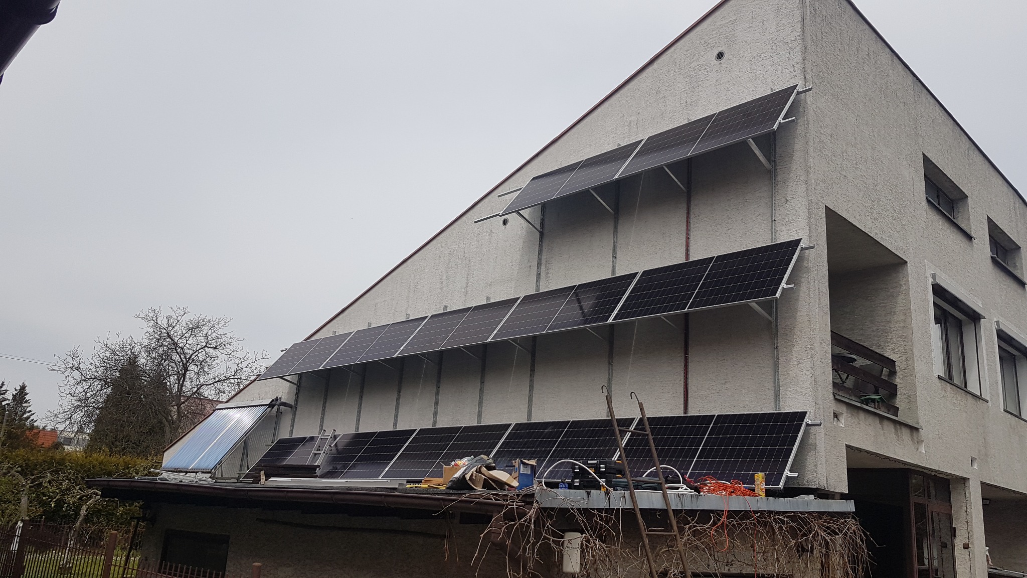 Montaż paneli słonecznych na elewacji budynku z widocznym dachem garażu i rozłożonymi narzędziami, pochmurne niebo w tle.