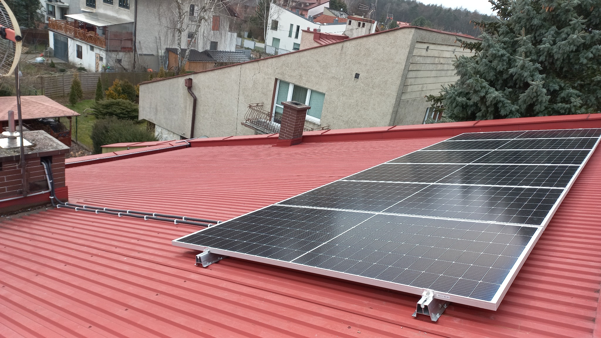 Panele słoneczne zamontowane na czerwonym dachu z blachy falistej, widoczne przewody prowadzone wzdłuż komina i dachu, w tle zabudowa jednorodzinna.
