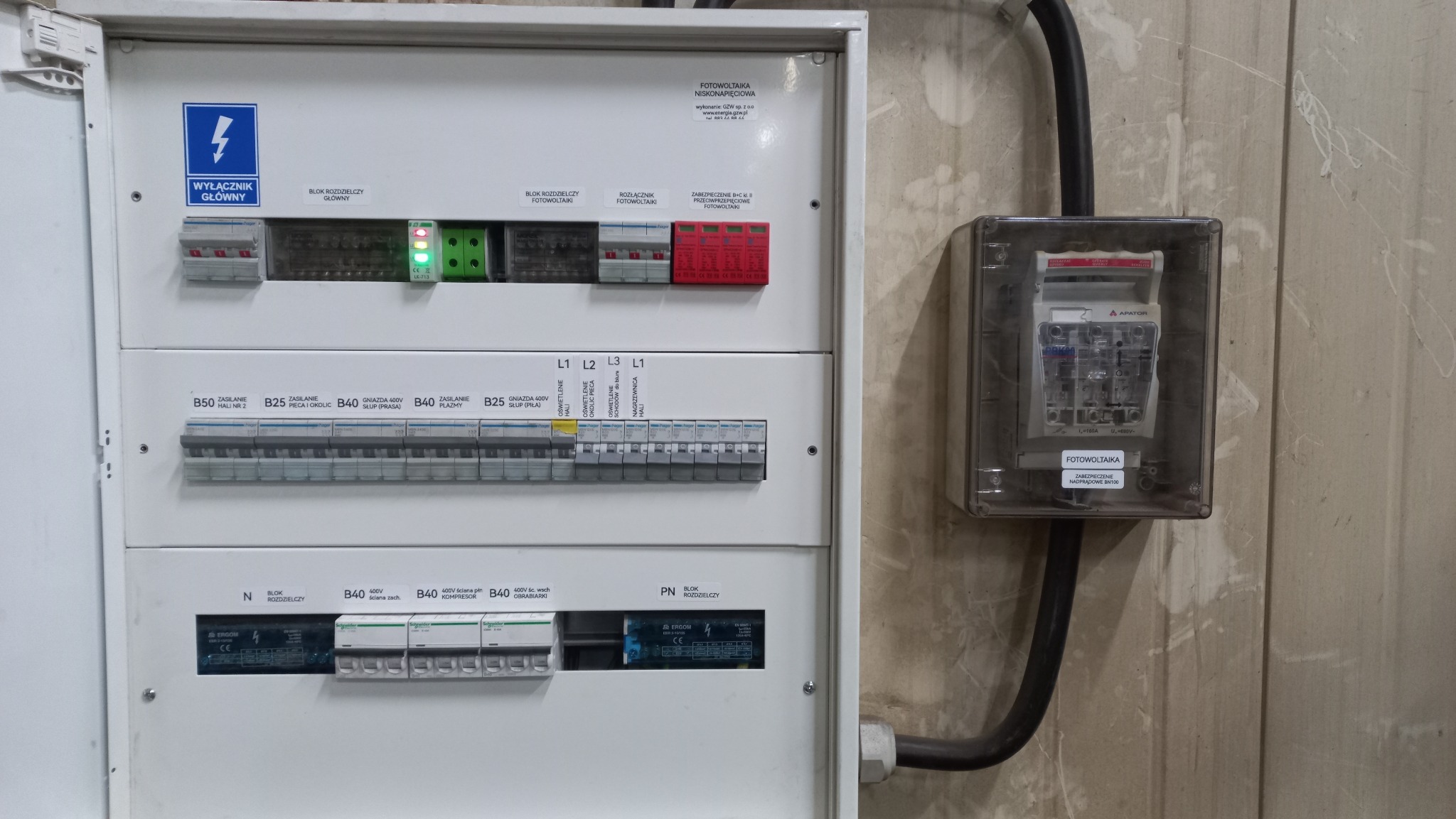Rozdzielnica elektryczna z zabezpieczeniami fotowoltaicznymi, widoczne wyłączniki, bloki rozdzielcze i zabezpieczenia przeciwprzepięciowe, etykiety opisujące poszczególne obwody i komponenty.