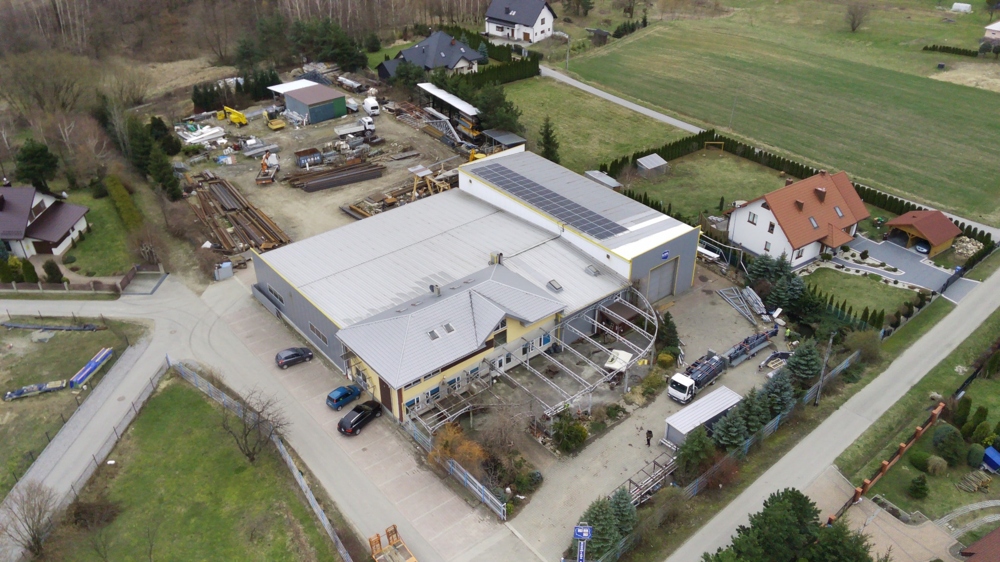 Widok z drona na budynek przemysłowy z panelami słonecznymi na dachu, składowisko materiałów budowlanych i teren budowy w otoczeniu domów jednorodzinnych.