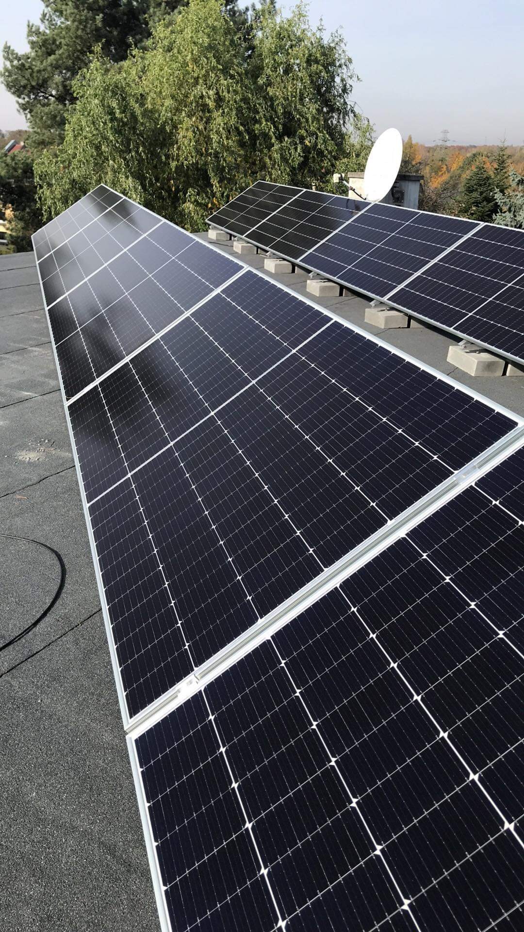 Panele słoneczne zamontowane na płaskim dachu z widoczną anteną satelitarną w tle.