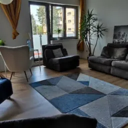 Jasne wnętrze pokoju z drewnianą podłogą, szarą sofą, fotelami w odcieniach szarości i niebieskiego, dywanem w geometryczne wzory oraz regałem z książkami i dekoracjami.