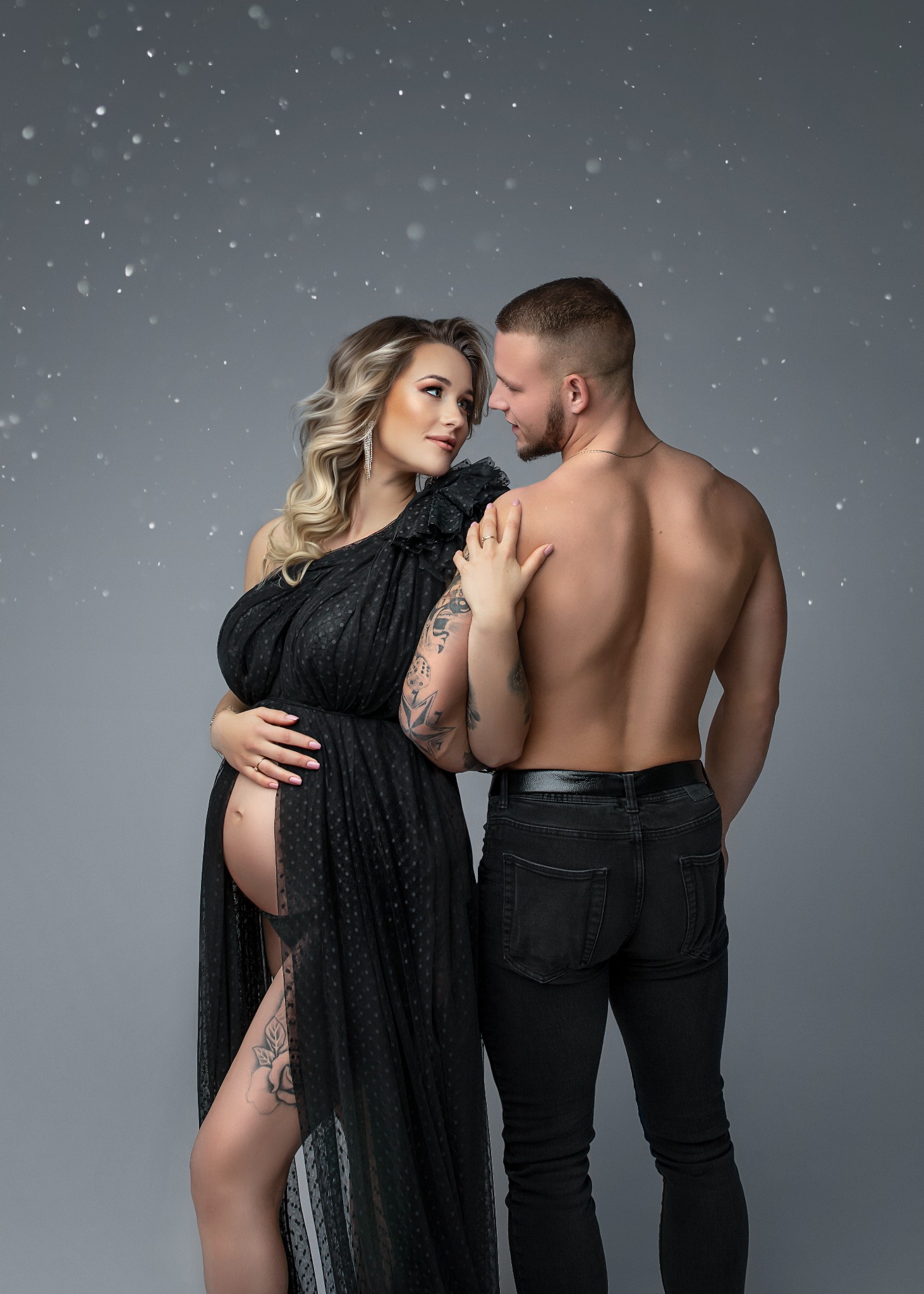 Kobieta w ciąży w czarnej sukni z odkrytym brzuchem i tatuażem na udzie, obejmowana przez mężczyznę bez koszuli na tle szarego studia z efektem brokatu.
