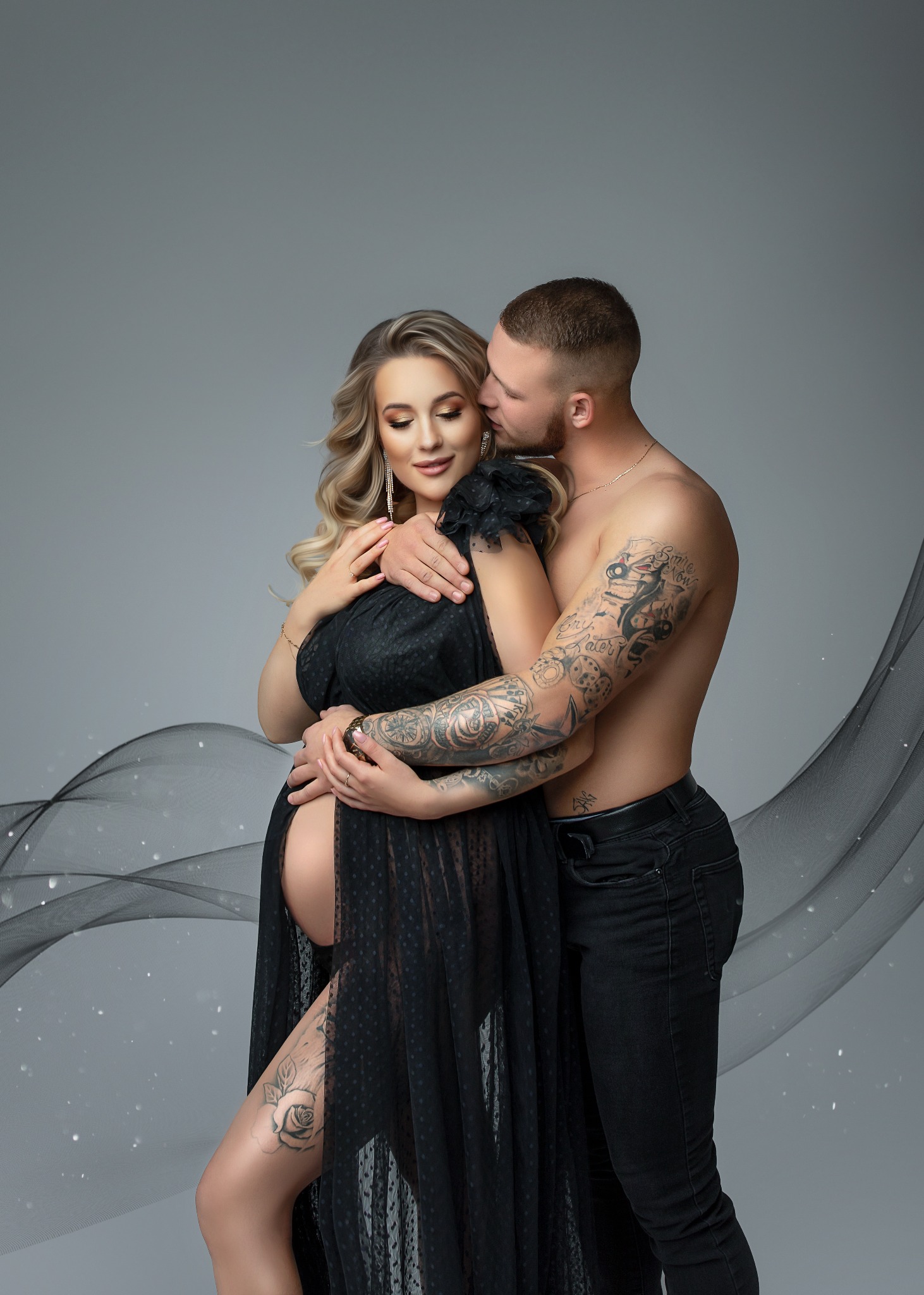 Elegancka, ciężarna kobieta w czarnej, prześwitującej sukni, obejmowana przez wytatuowanego mężczyznę na szarym tle z subtelną dekoracją z tiulu.