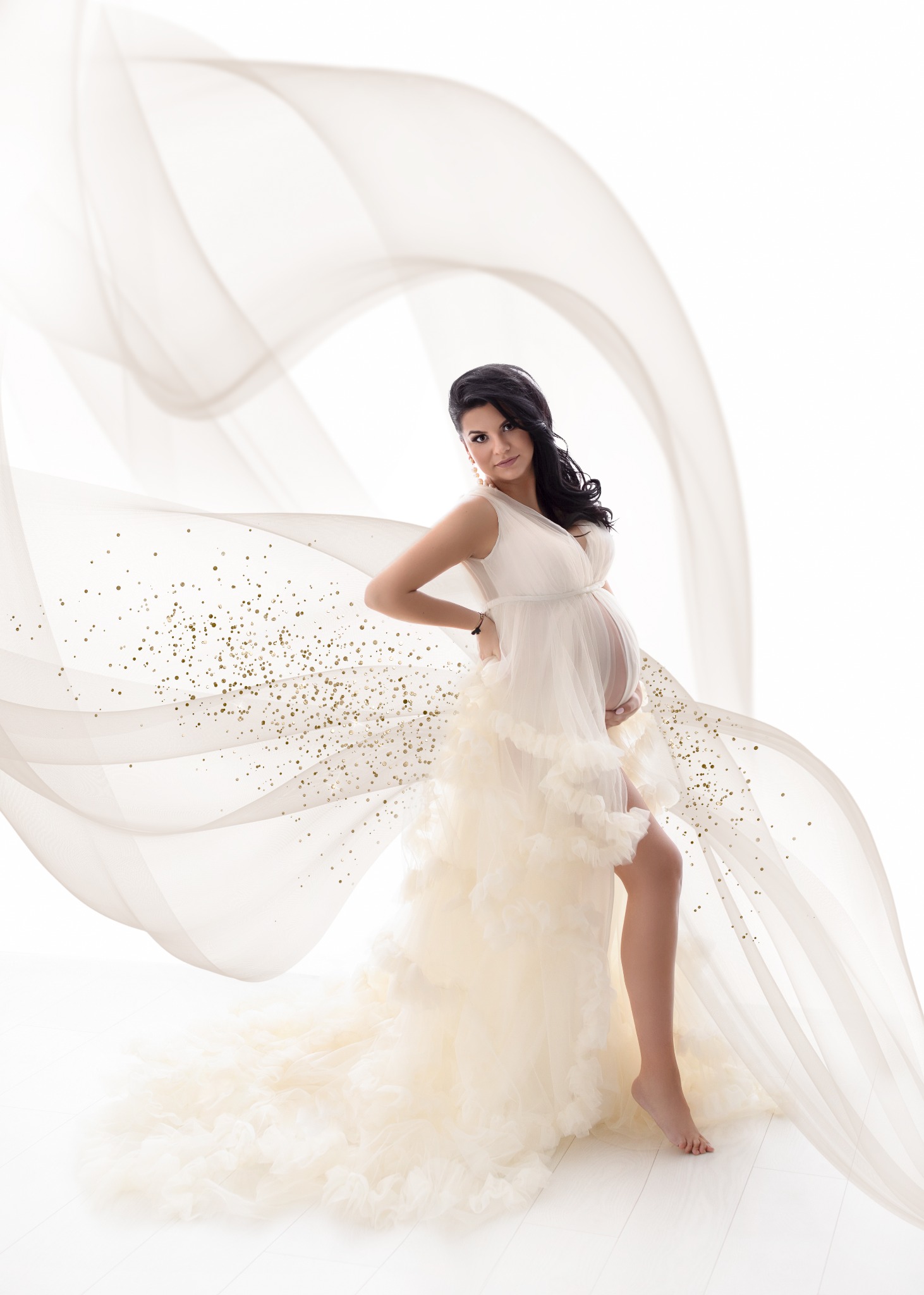 Kobieta w ciąży w zwiewnej, kremowej sukni z falbanami, pozująca na białym tle z powiewającą tkaniną ozdobioną złotymi drobinkami.