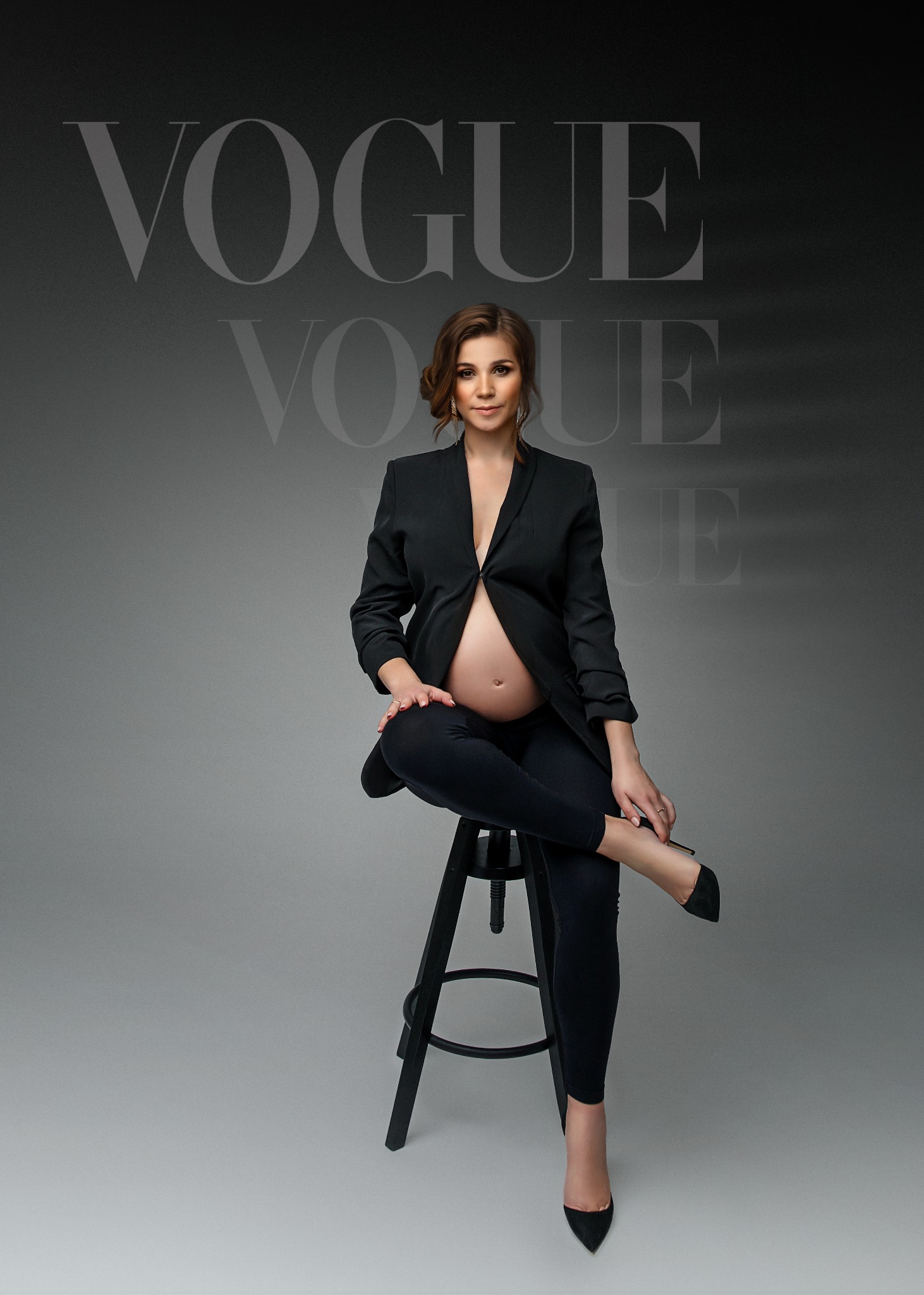 Kobieta w ciąży w czarnym garniturze siedzi na stołku na szarym tle z napisem VOGUE.
